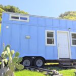 Blue tiny house on wheels with small windows and white door in a grassy area.