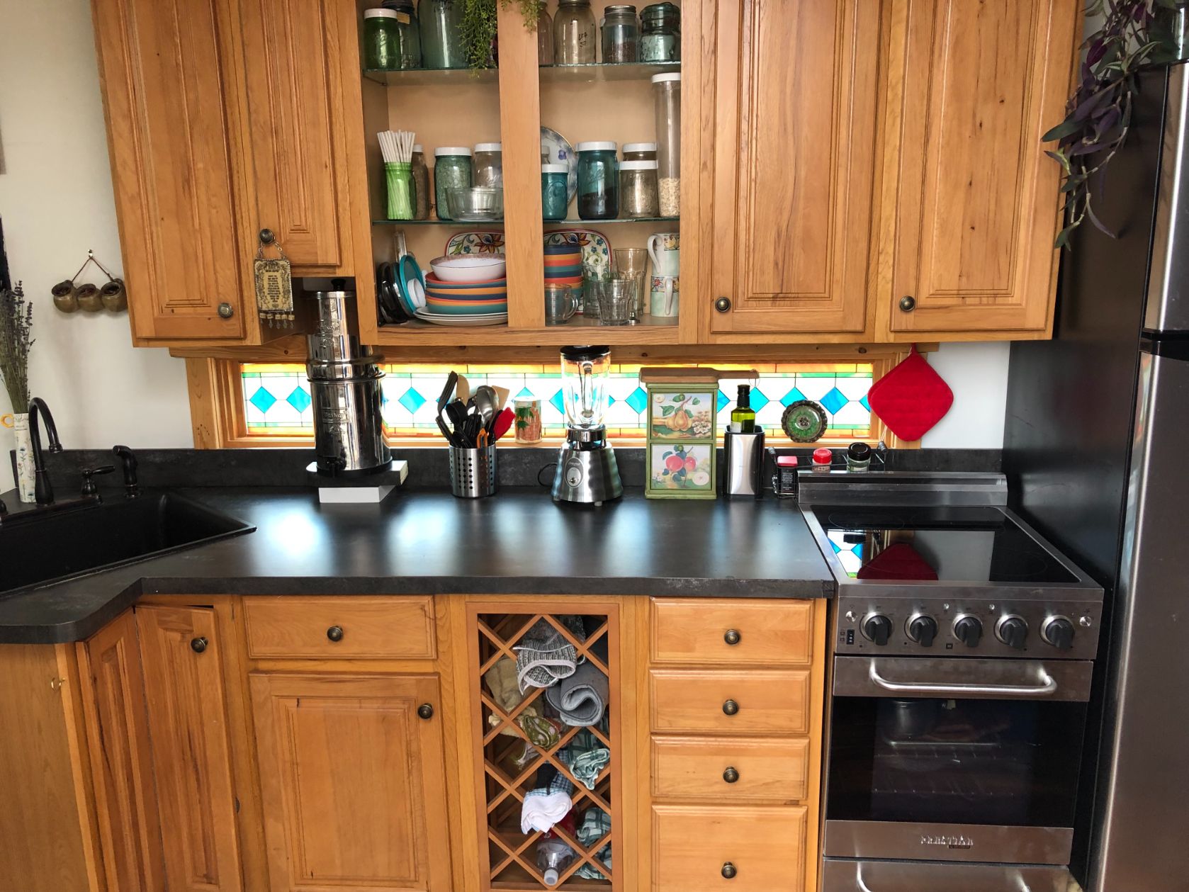 Kitchen area of the Kangaroo THOW featuring stained glass backsplash window and honey-colored cabinetry