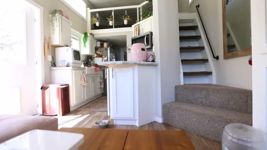 Tiny house interior with white kitchen cabinets, island and carpeted staircase to the loft