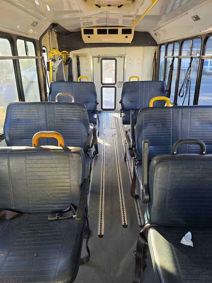 Interior aisle with dark vinyl bus seats, yellow grab bars, and floor tie-down tracks