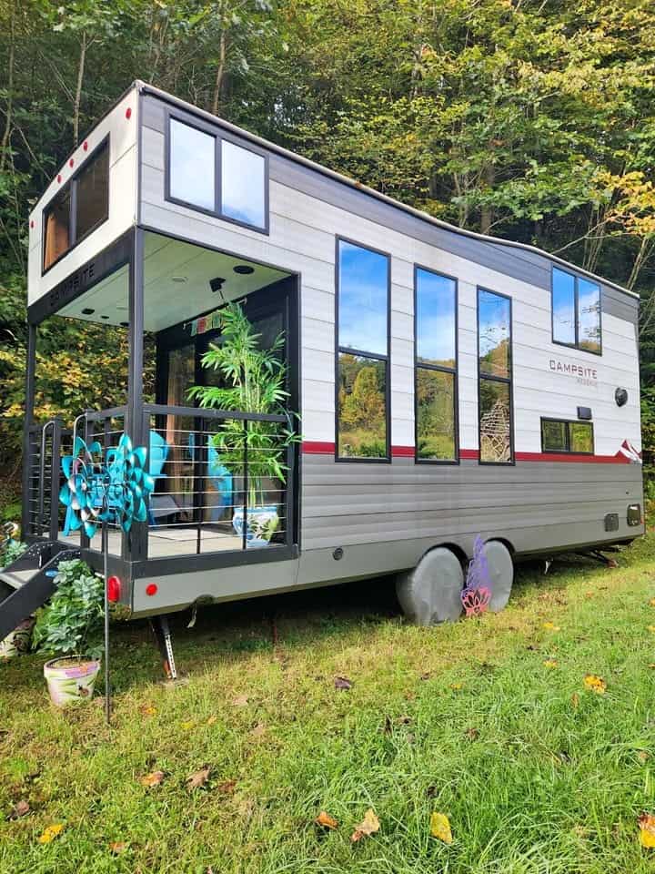Gray and white Forest River Campsite Reserve tiny house trailer with tall windows and a small front porch on a grassy wooded lot