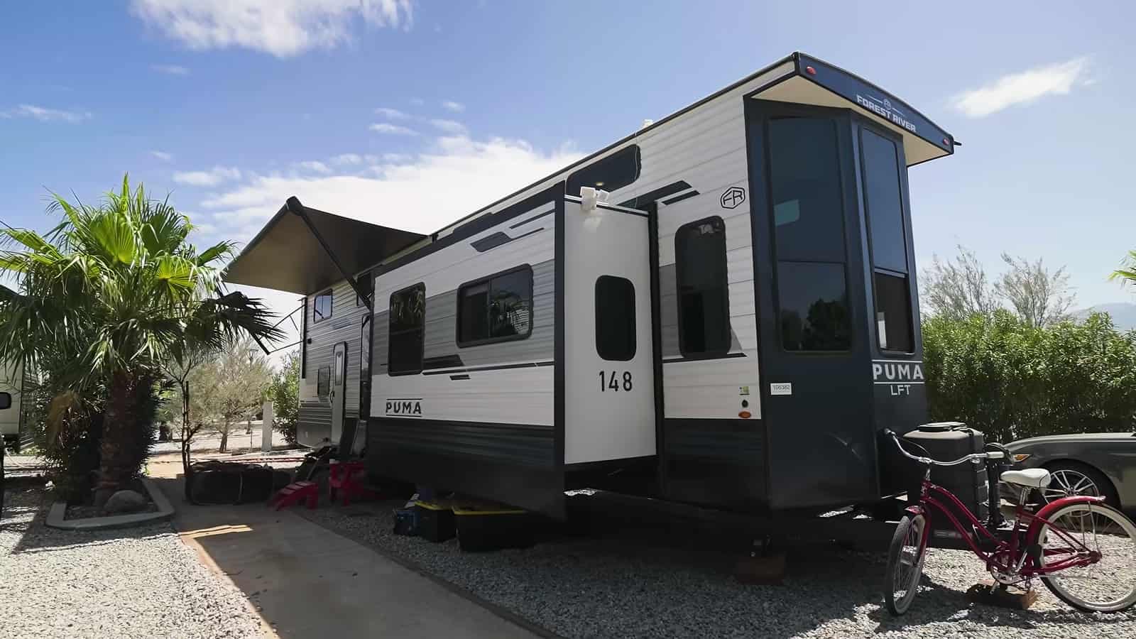 Forest River PUMA destination trailer with slide-outs and an awning on a gravel lot beside a palm tree and a red bicycle