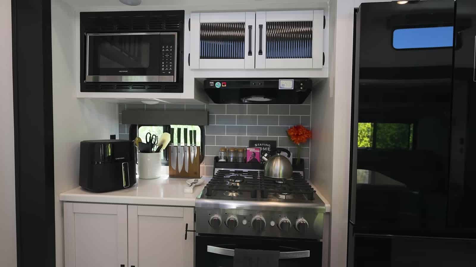 Gas range with a hood, microwave, glass-front cabinets, and gray subway tile backsplash in the trailer kitchen