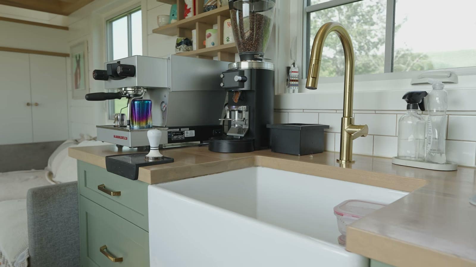Espresso machine and grinder beside a white farmhouse sink with a brass faucet and sage-green cabinets