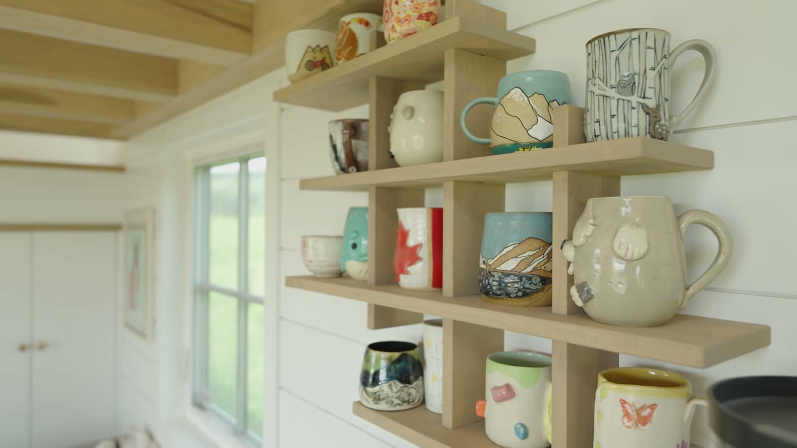 Open wooden shelves filled with handmade pottery mugs next to a window