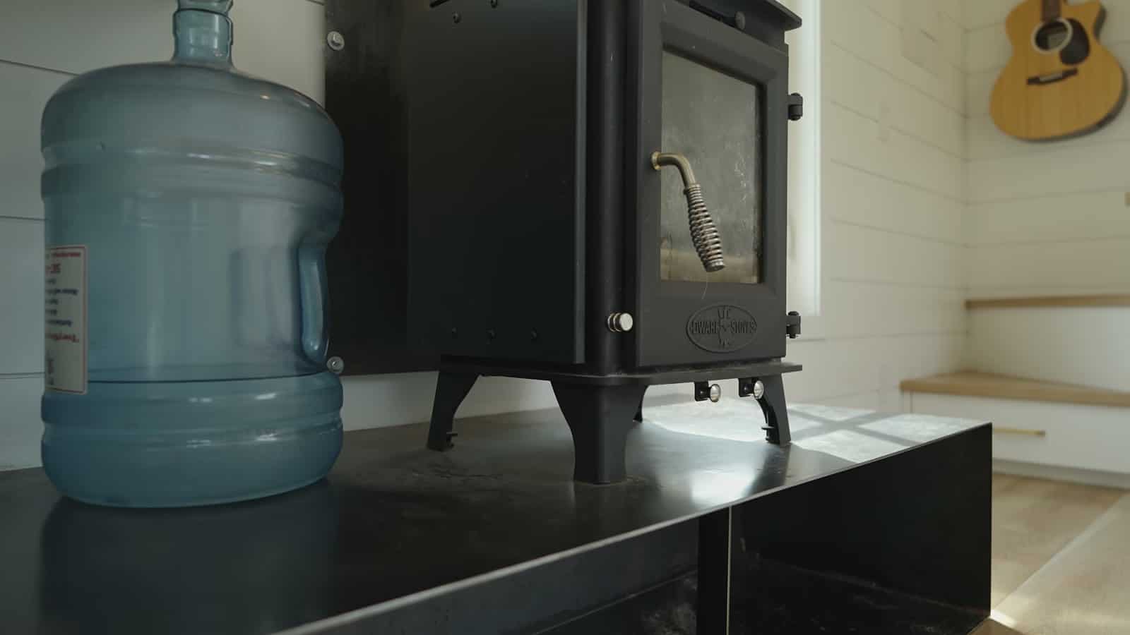 Small black wood stove on a raised hearth with a water jug and an acoustic guitar on the wall