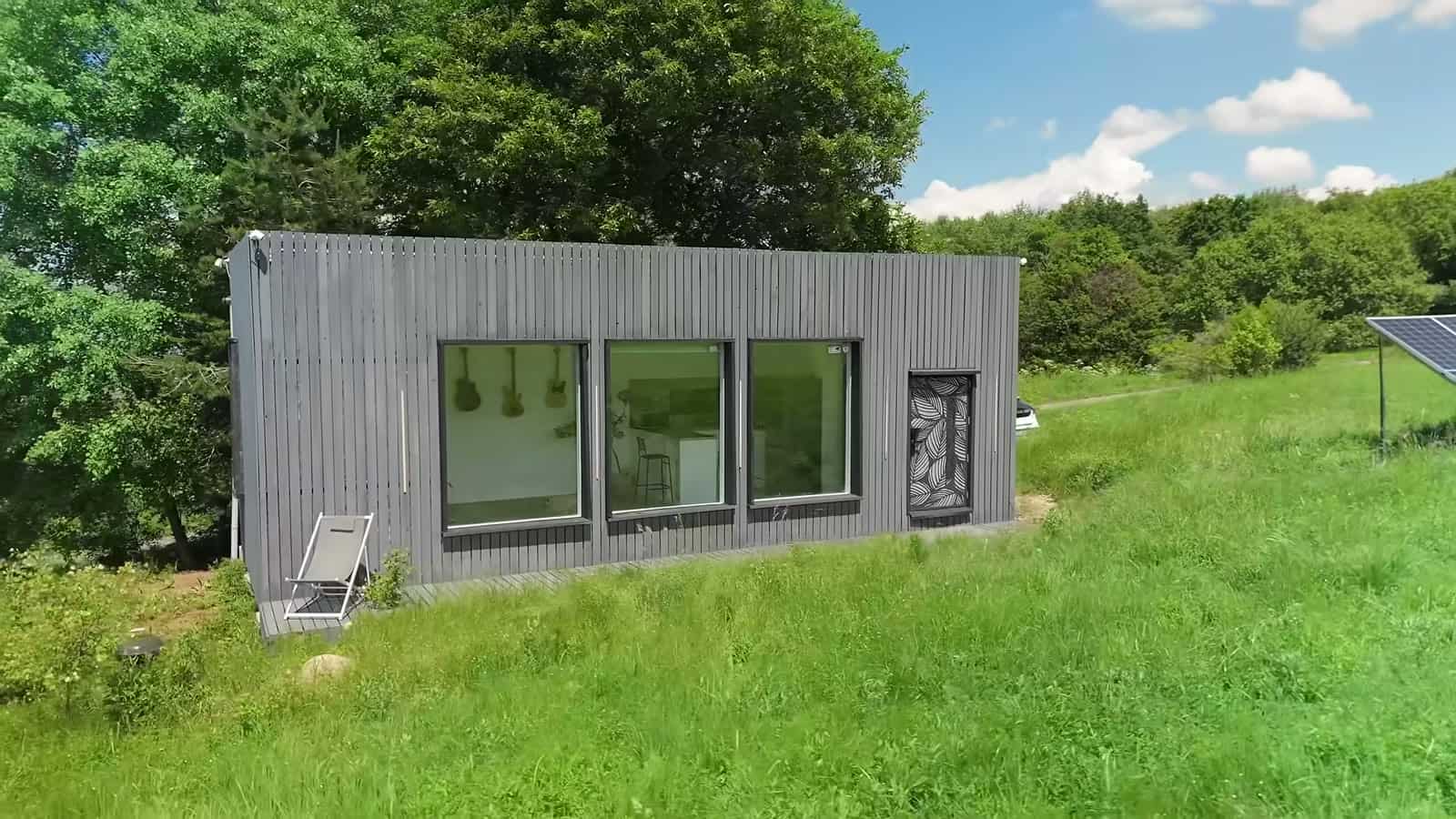 Gray wood-clad off-grid studio with three large windows in a grassy meadow, with a solar panel at the edge of the lawn