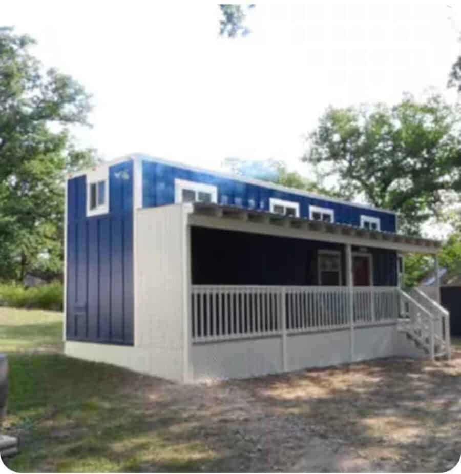Blue and white tiny house on wheels with a long covered front porch, seen from the side on a grassy lot