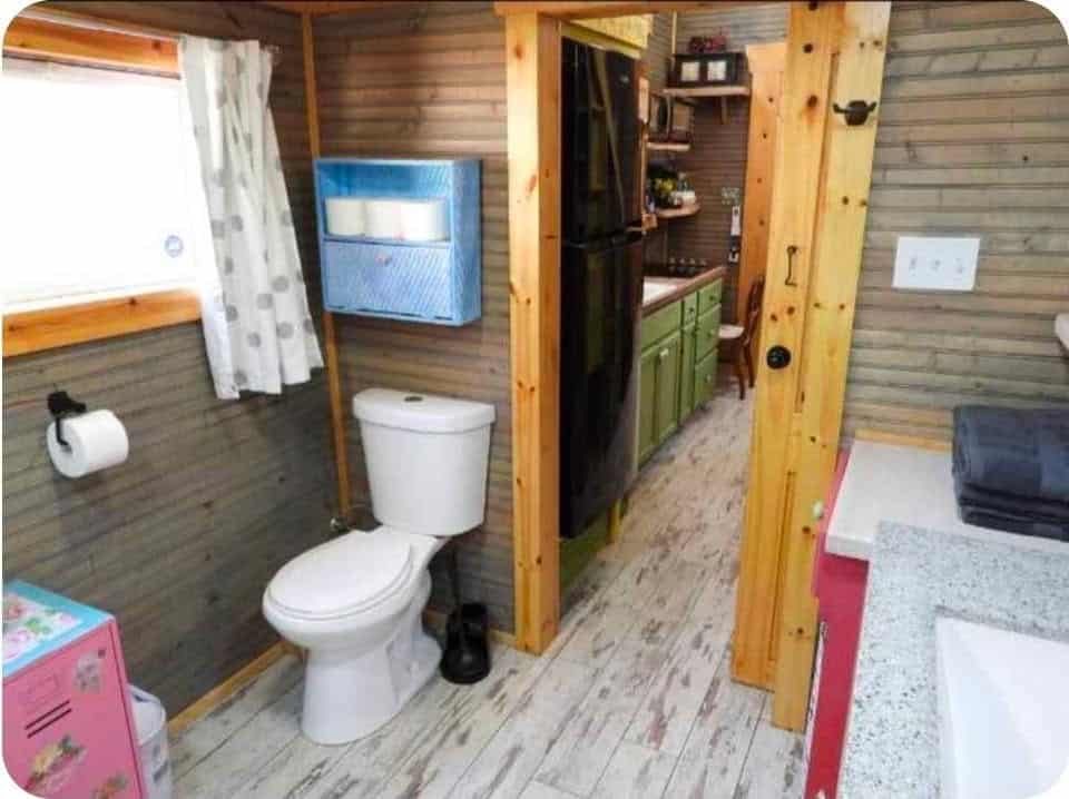 View from the bathroom toward the kitchen, with a toilet, wall heater, and pine trim