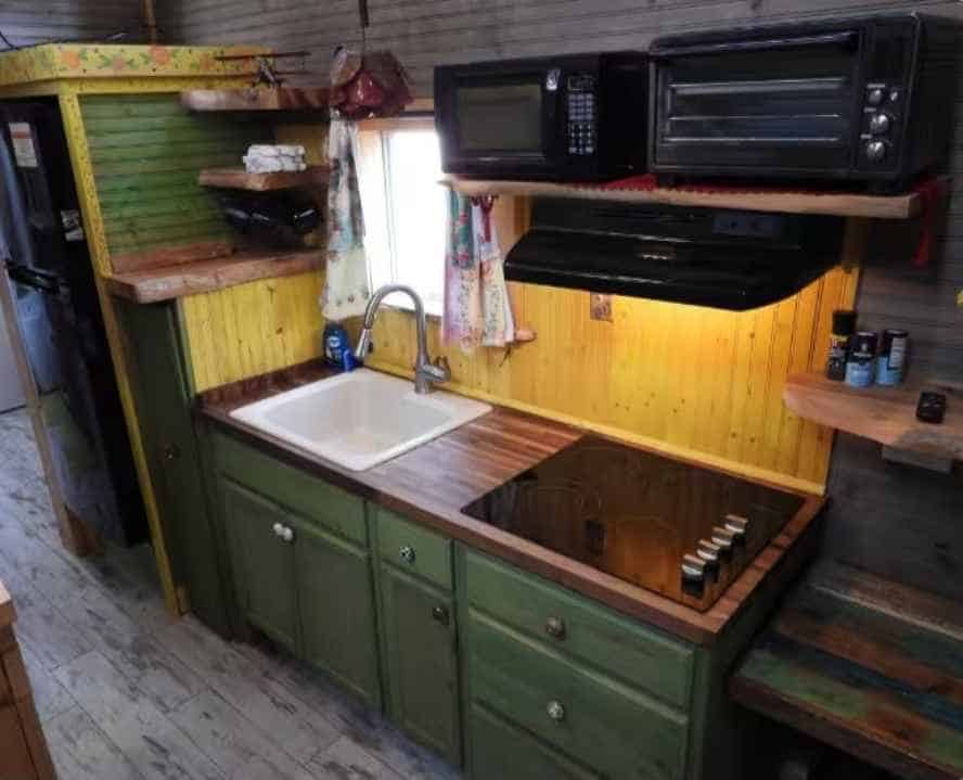 Tiny house kitchen with green lower cabinets, butcher block counter, white sink, induction cooktop, microwave, and countertop oven