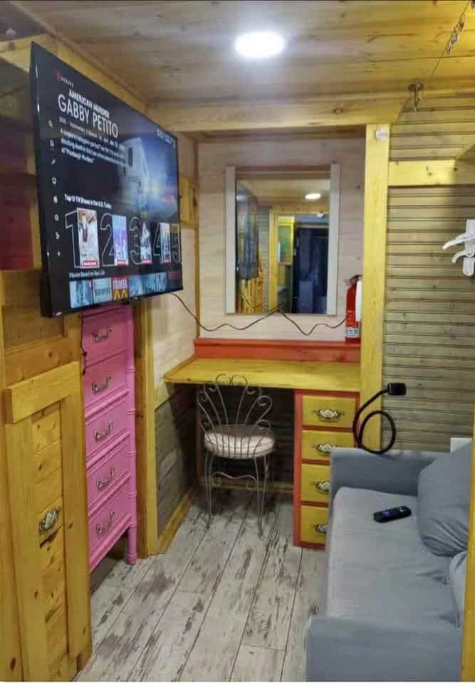 Living area with a wall-mounted TV, pink dresser, built-in desk and vanity mirror, and a gray sofa under a wood ceiling