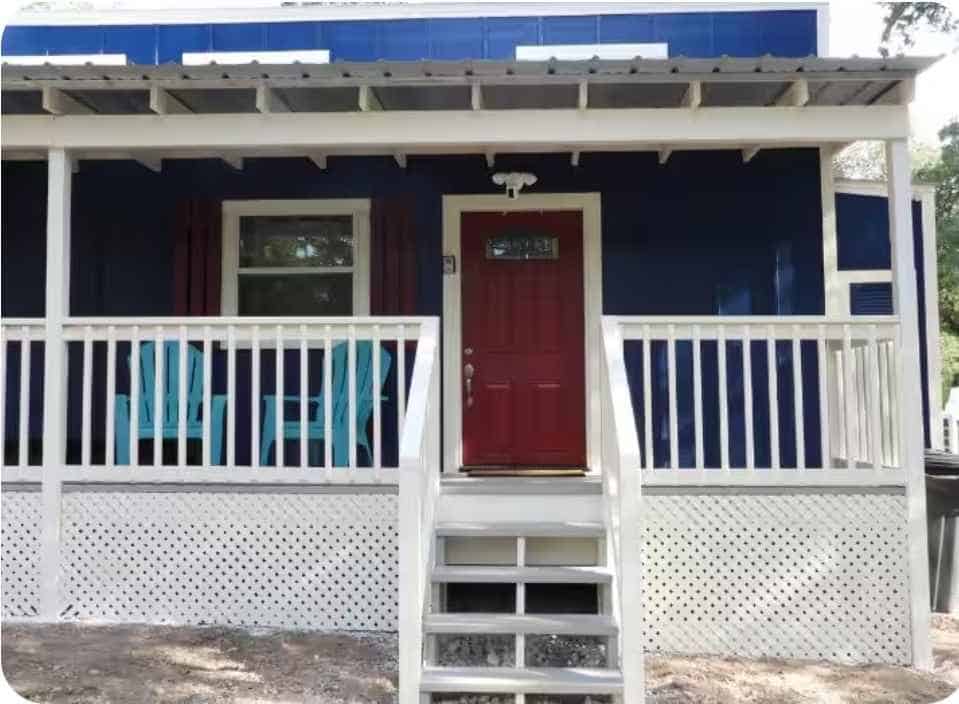 Front of the tiny house: red entry door, white porch railing, lattice skirting, and wooden stairs
