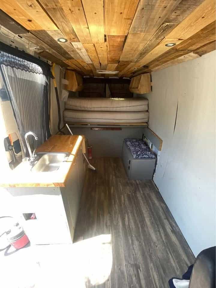 Inside the Sprinter: reclaimed wood ceiling, galley counter with sink, wood-look floor, and the bed area at the back