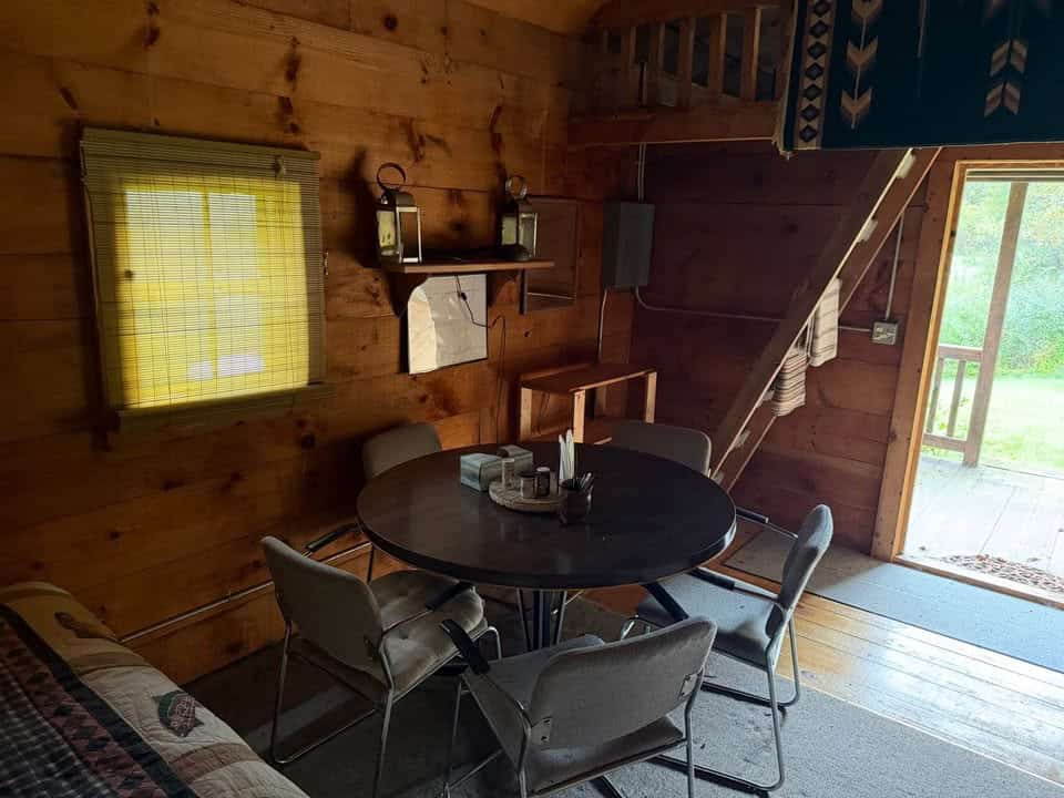 Round dining table and chairs below the loft ladder, with log walls and a window