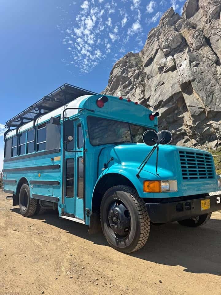 Teal 1994 Ward shortbus with a black roof rack parked on a dirt lot beside a rock outcrop