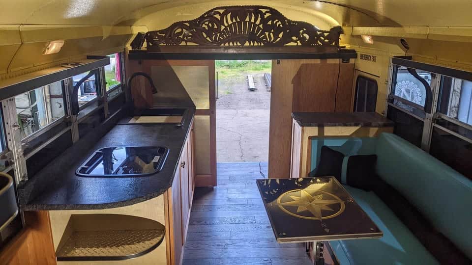 Inside the shortbus: granite counters, wood cabinets, a carved wood valance, and a teal bench dinette with a compass-inlay table
