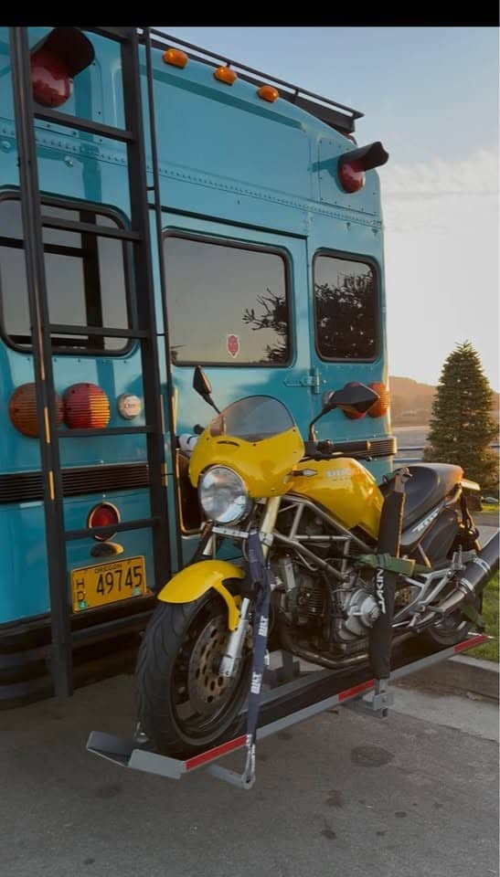 Rear of the bus with a ladder to the roof rack and a yellow motorcycle on a hitch carrier