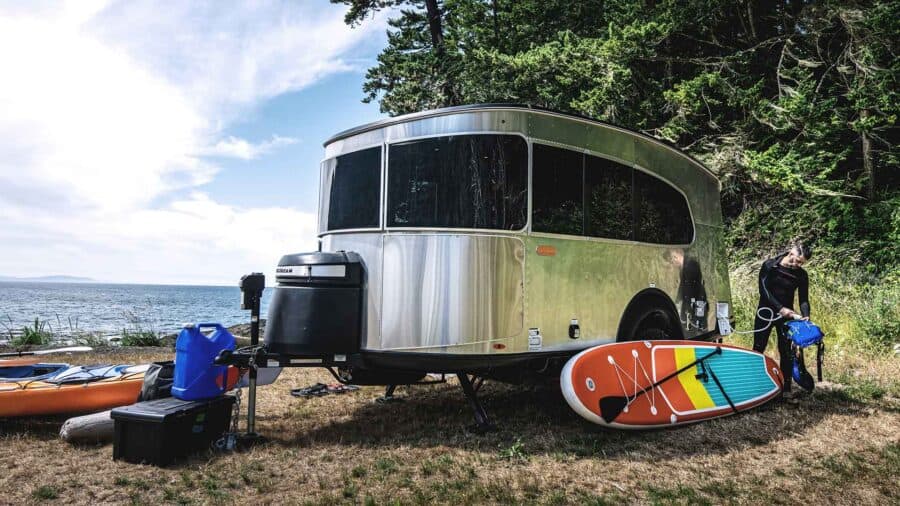 Silver 2027 Airstream Basecamp single-axle travel trailer on dry grass at the water, wraparound front window, black tongue pod, paddleboard against the side, kayak on the bank, person at the rear with a hose