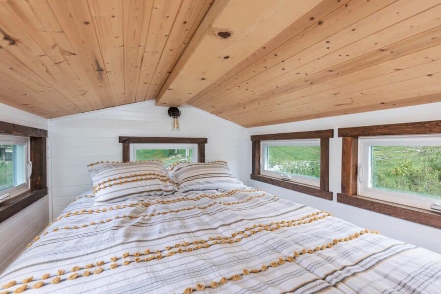 Interior of a tiny home bedroom with a sloped wooden ceiling and multiple windows for natural light.