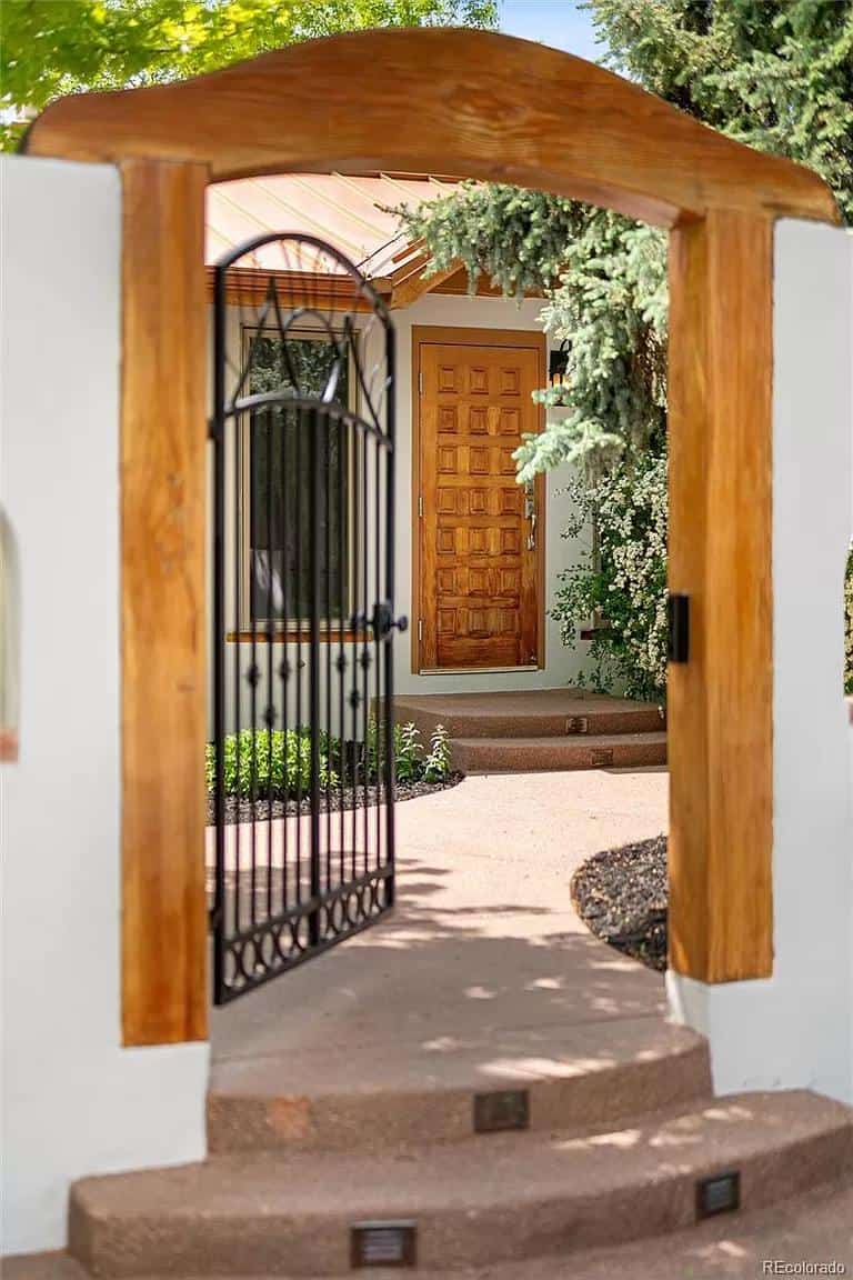 Front view of a charming Spanish-style tiny house with a wooden gate and lush greenery.