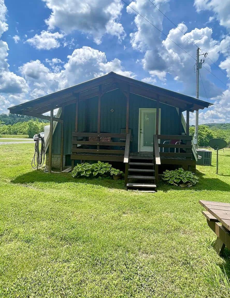 A-Frame tiny house with metal roof and siding, set in a lush green landscape under a bright blue sky.