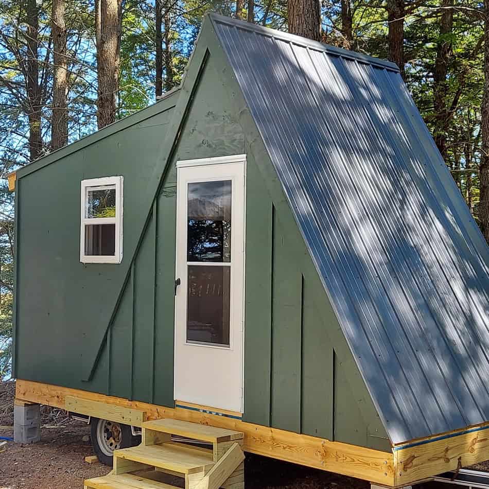 A-Frame tiny house with green exterior and metal roof in wooded setting.
