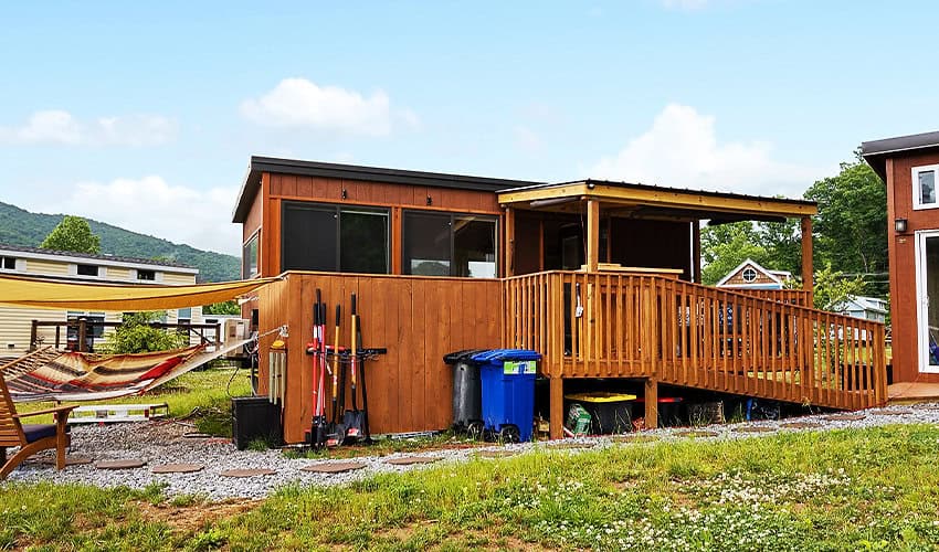 Small wooden tiny house with large deck and outdoor gear, set in a green landscape.