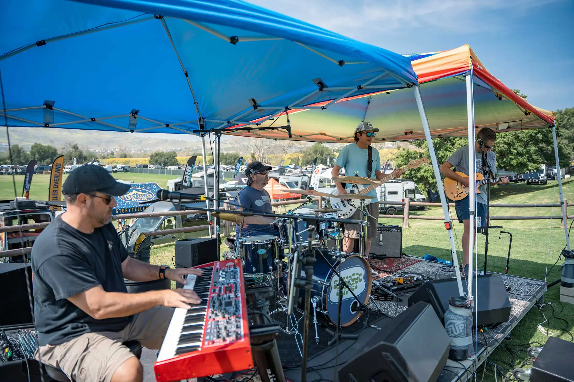Four piece band playing on a small stage under a canopy with vans parked in the field behind