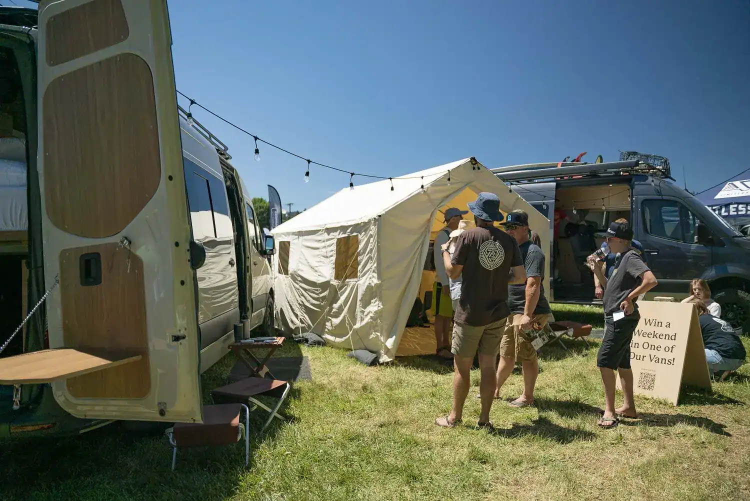 Open Sprinter van with wood interior beside a canvas wall tent and attendees at an outdoor van expo