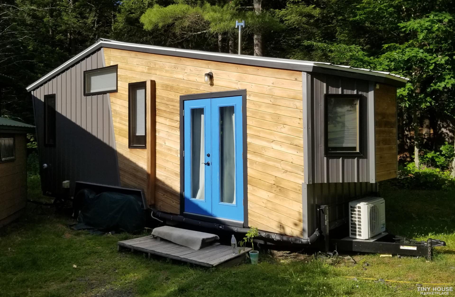 Small modern tiny house with blue door, wooden exterior, and large windows in a green setting.