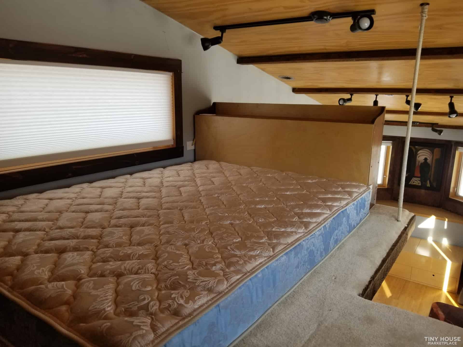 Sleeping loft with full mattress wood plank ceiling track lighting and window with honeycomb blind