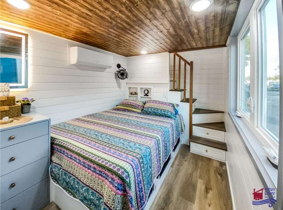 Compact tiny house bedroom with built-in stairs and large windows.