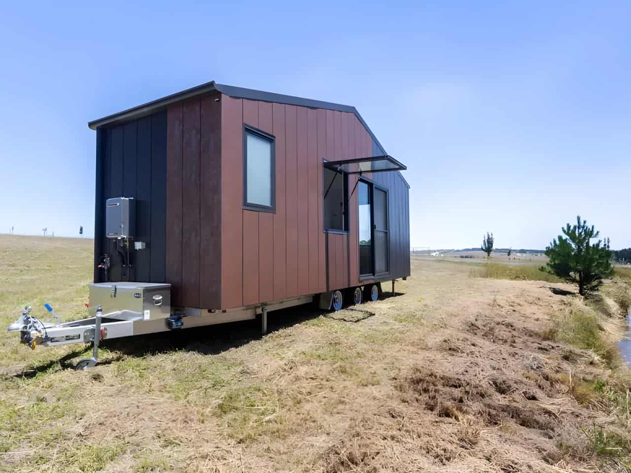 Rear three-quarter view of the Audrey tiny house showing the two-tone aluminium and timber-look cladding, external gas hot water unit, propane storage box on the drawbar, and awning servery window