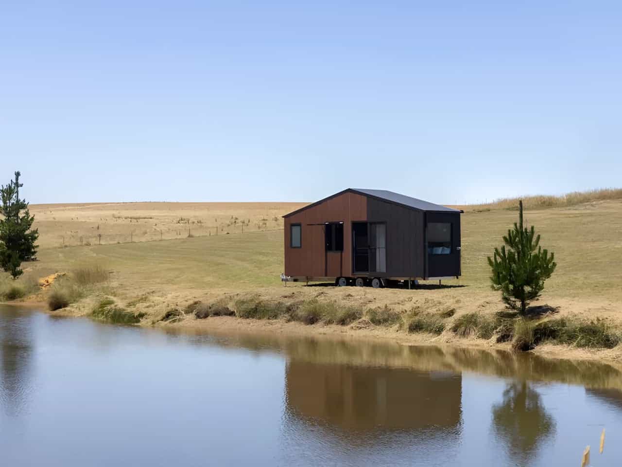 Wide view of the Audrey tiny house sited on the bank of a dam in open Australian farmland, showing its compact single-level profile and gabled roofline