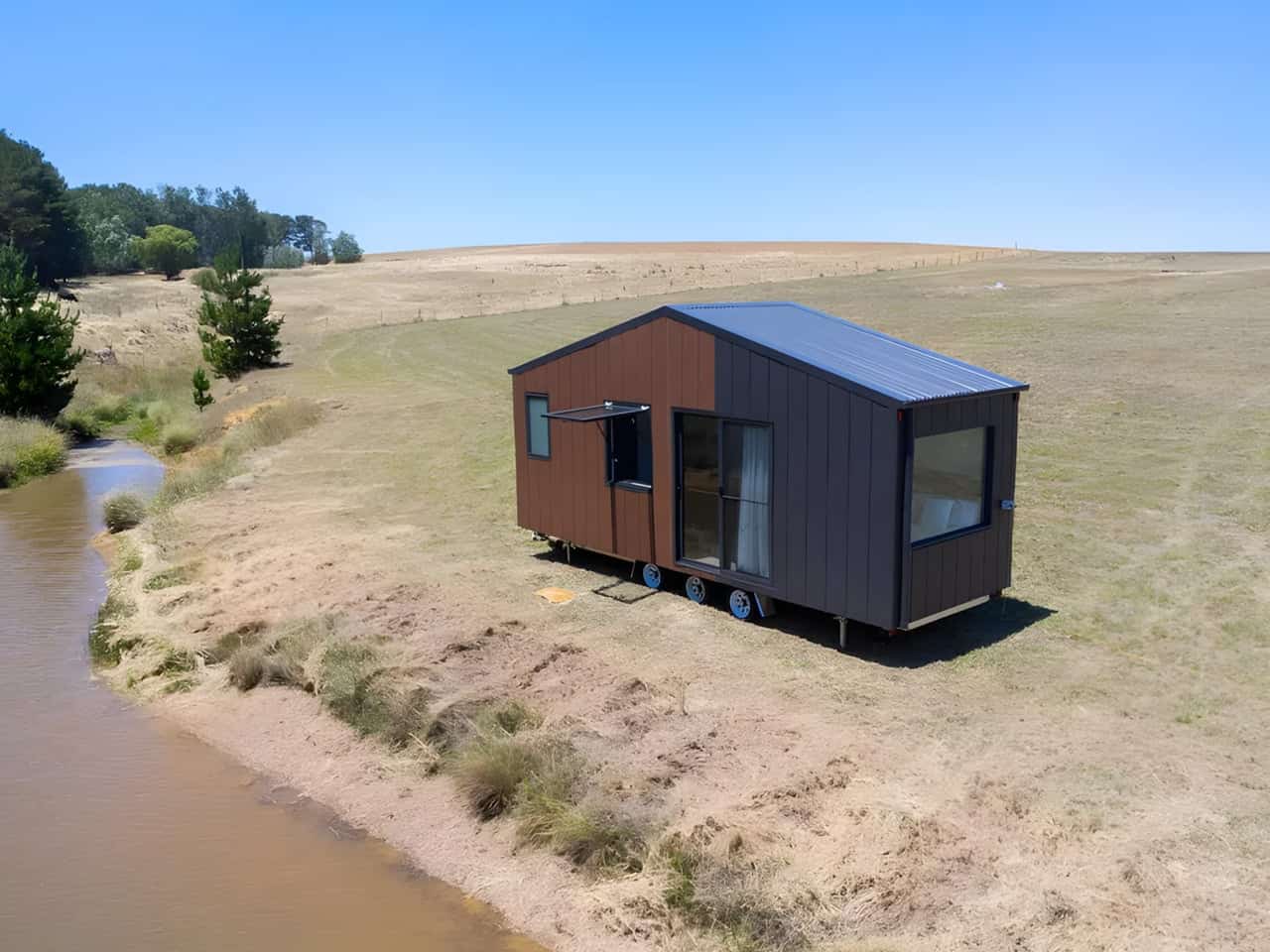 Audrey tiny house by CozyCo with corrugated aluminium and timber-look cladding, dark metal roof, and corner picture window, parked beside a dam on rural Australian farmland