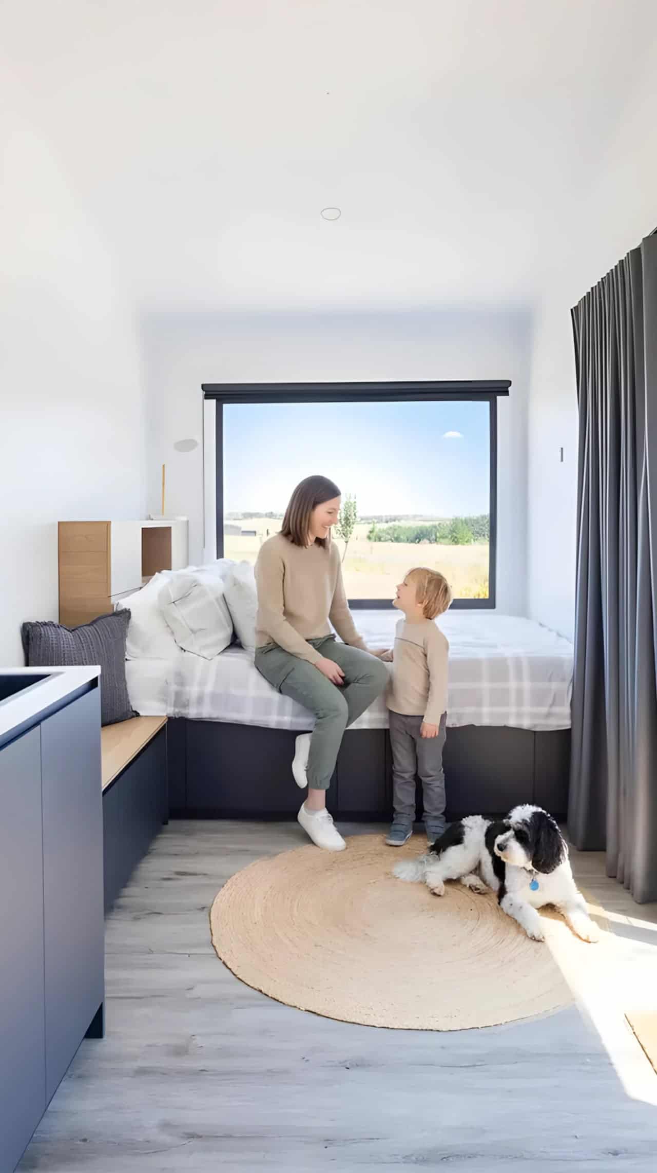 Open-plan interior of the Audrey tiny house with white walls, pitched ceiling, light timber-look flooring, navy kitchen joinery, and the storage bed beneath a picture window