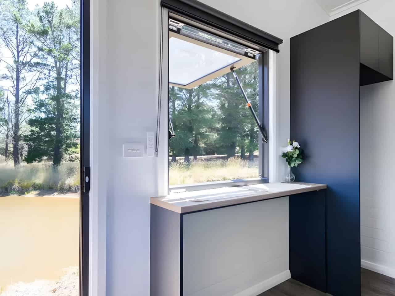 Interior of the Audrey tiny house showing the kitchen bench beneath a wide awning servery window propped open to the outdoors, with navy cabinetry and white VJ paneling