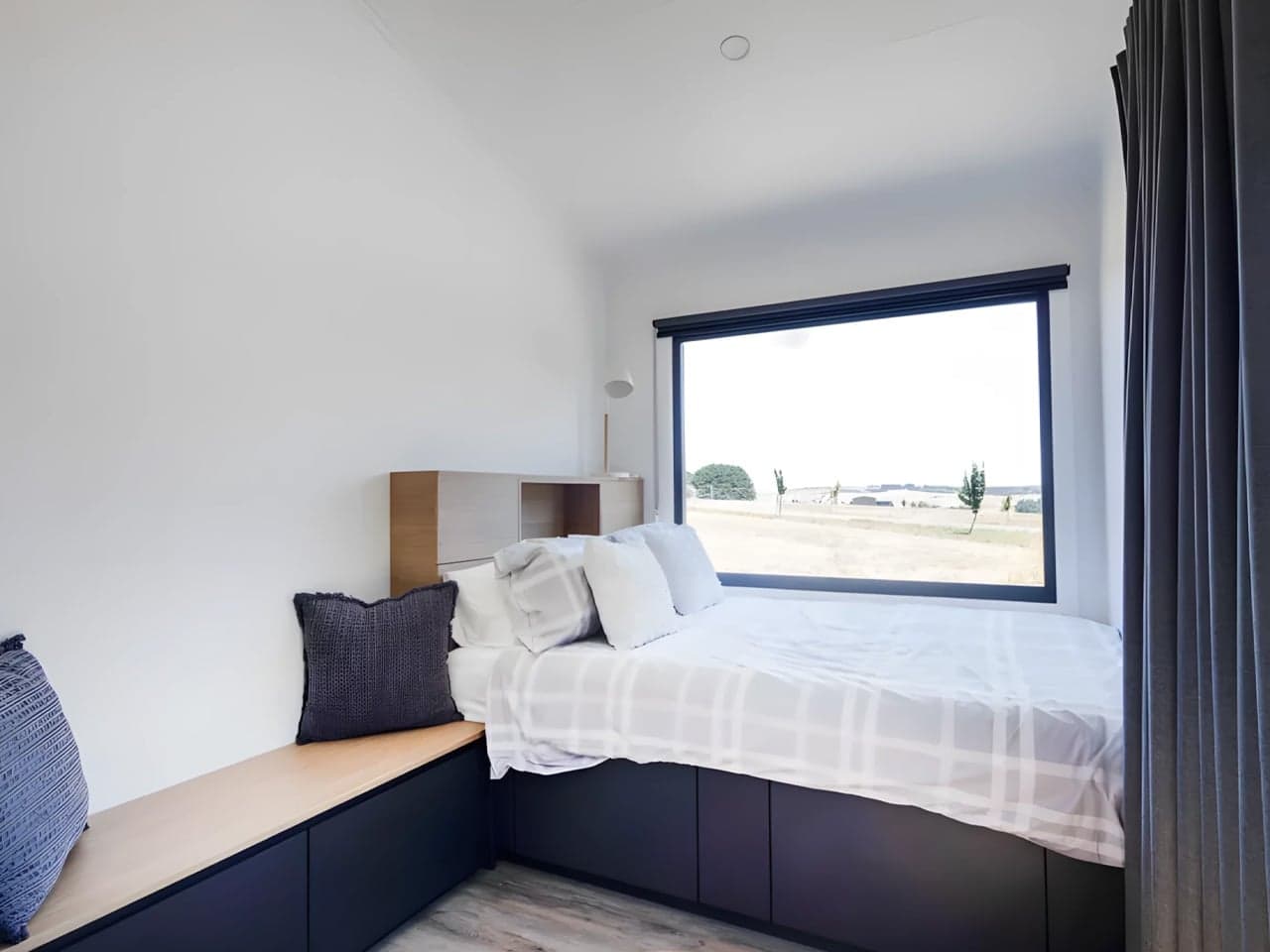 Raised storage bed in the Audrey tiny house set beneath a large picture window, with navy drawer fronts in the base, a timber-topped bench seat, and a built-in headboard shelving unit