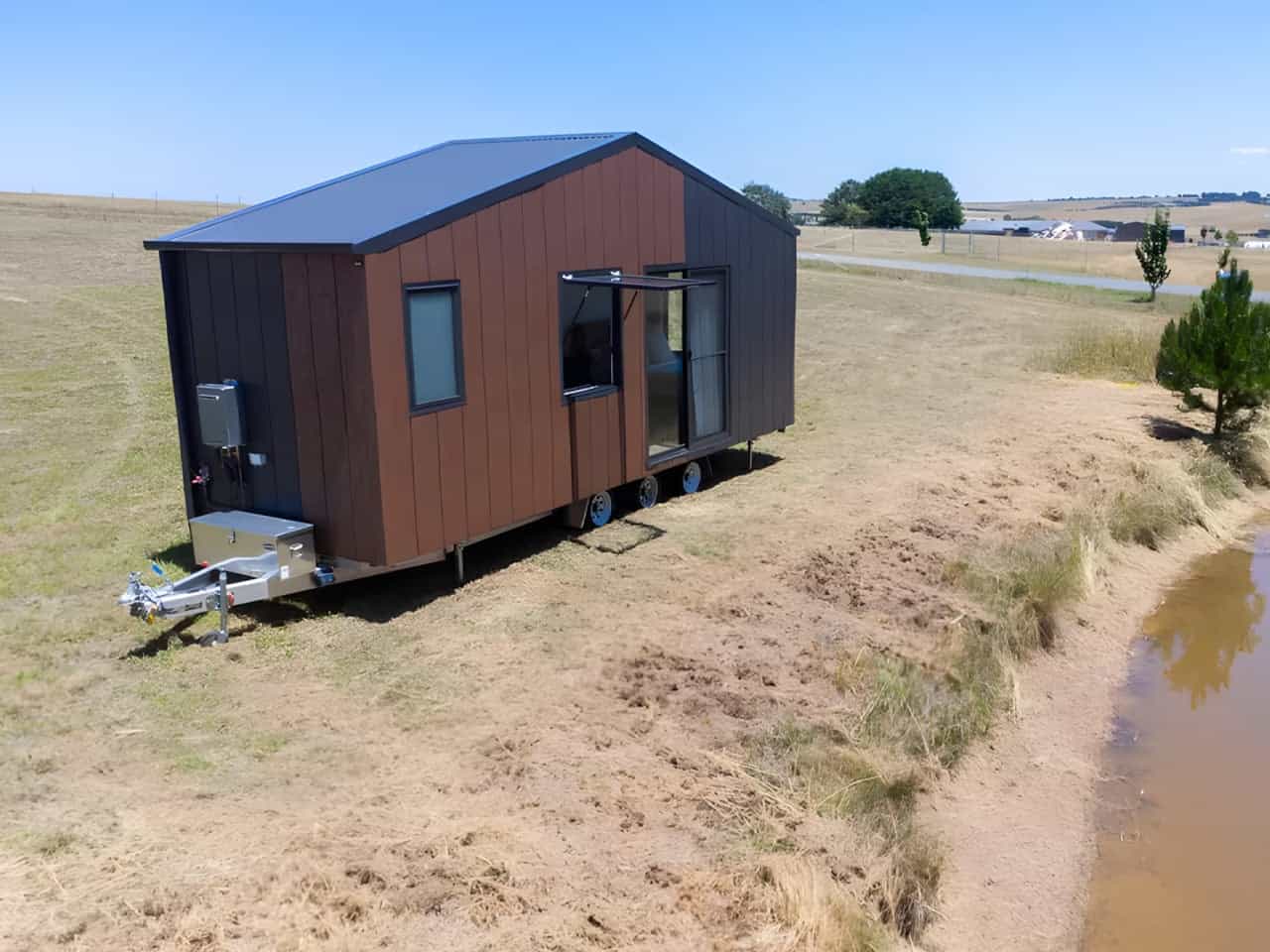 Aerial view of the Audrey tiny house showing its triple-axle trailer, drawbar-mounted storage box, external gas hot water system, and drop-down stabilizing legs