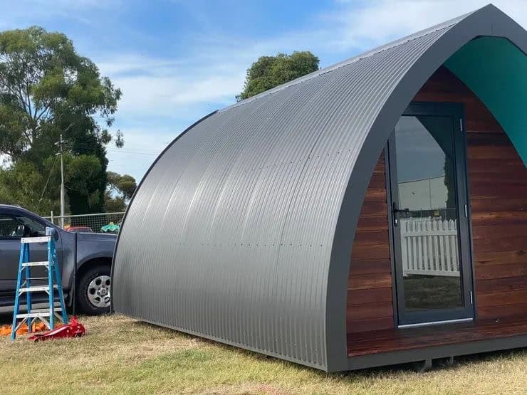 Arched corrugated steel cabin with timber cladding and glass door on display at an expo