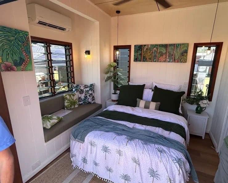 Bedroom interior of an Australian tiny home with window seat, tropical artwork, and air conditioning