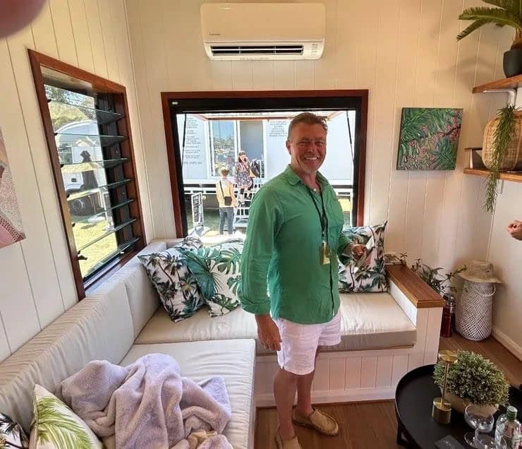 Builder standing inside a tiny home lounge with a window seat, cushions, and split system air conditioner