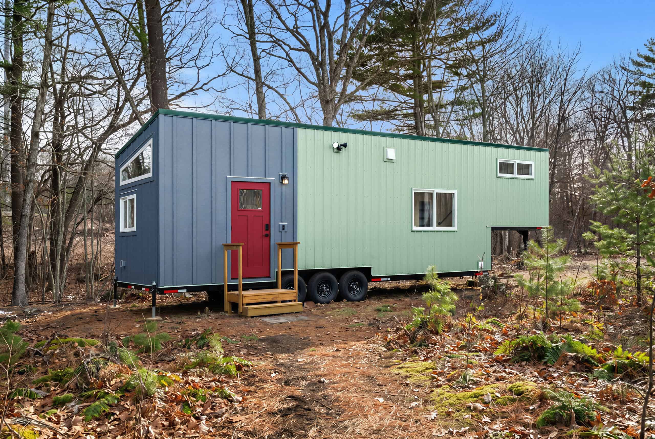 The Knoll by Backcountry Tiny Homes, a 38-foot gooseneck tiny house with two-tone metal and board-and-batten siding and a red door, parked in the woods