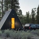 A-frame tiny house on wheels with black exterior in a forest setting.