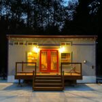 Wide tiny home on wheels with large front deck and bright interior lighting.