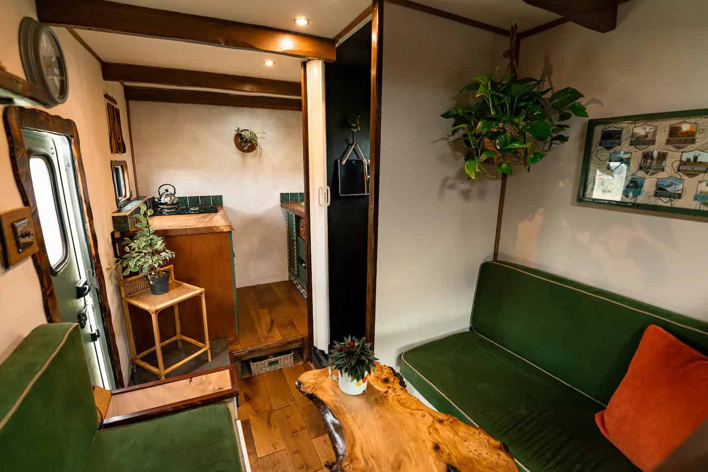 1970s-style van interior with green corduroy seating, live-edge table, hanging plant, and a galley kitchen beyond