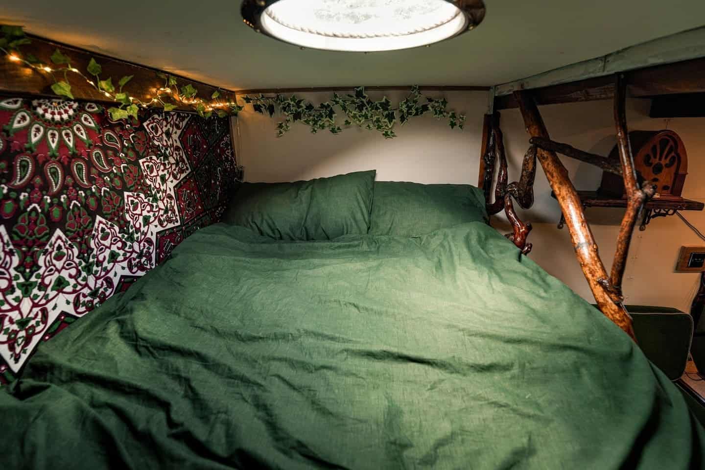 Upper double bed under a glowing skylight dome with tapestry, ivy, fairy lights, and a driftwood bed divider