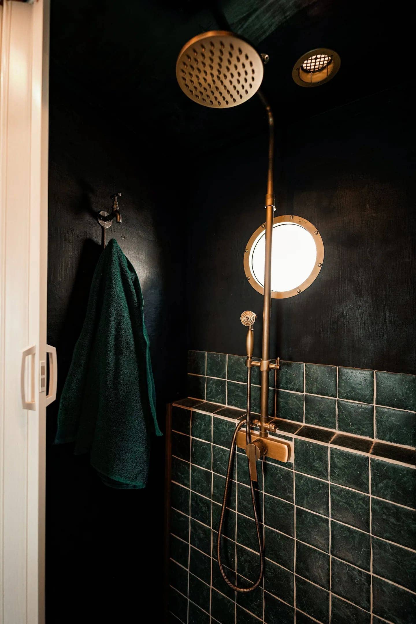 Green-tiled van wet room with a brass rainfall shower head, brass porthole window, and dark textured walls