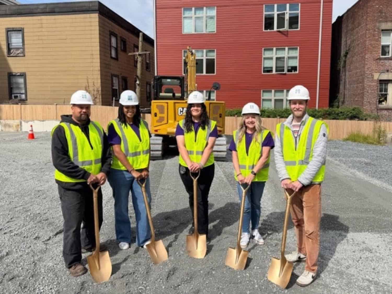 Five people in hard hats and safety vests holding gold shovels at the Belmont Tiny House Village groundbreaking on a gravel lot in Capitol Hill Seattle