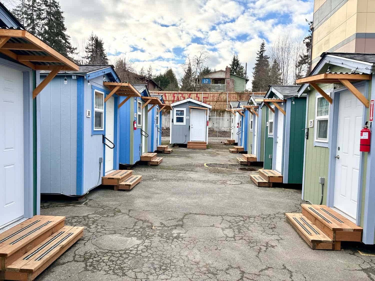 A row of blue and green wood-framed tiny houses with shingled gable roofs, cedar porch awnings, numbered doors and wooden steps at a Seattle tiny house village